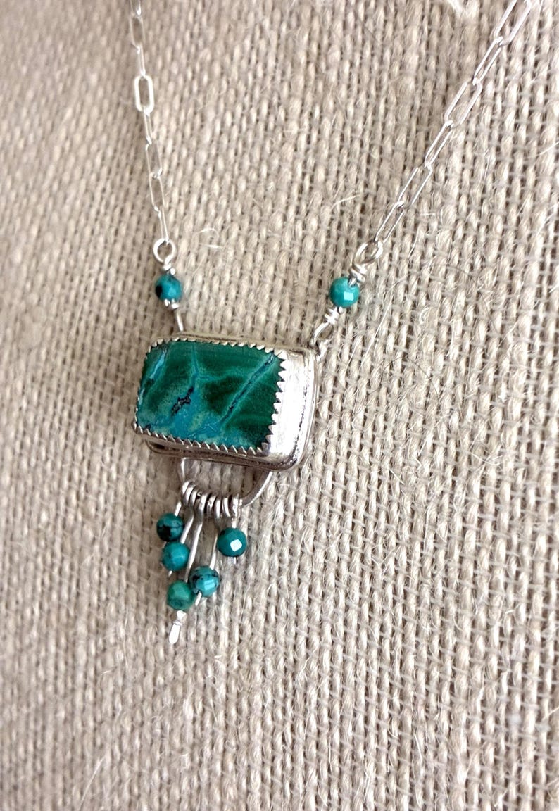 May include: A silver necklace featuring a rectangular malachite pendant with a serrated bezel setting. The pendant is accented with small turquoise beads and a dangling fringe of silver wires tipped with additional turquoise beads.
