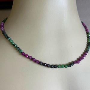 May include: A delicate necklace made of faceted green and pink gemstones. The necklace is strung on a thin black cord and features a graduated color scheme, with the green stones at the center and the pink stones at the ends.