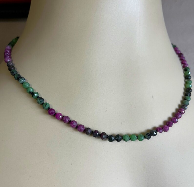 May include: A delicate necklace made of faceted green and pink gemstones. The necklace is strung on a thin black cord and features a graduated color scheme, with the green stones at the center and the pink stones at the ends.
