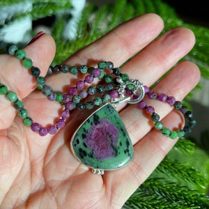 May include: A green and pink gemstone pendant necklace with a silver chain and clasp. The pendant is a teardrop shape and has a green and pink color pattern. The necklace is made of small green and pink beads.