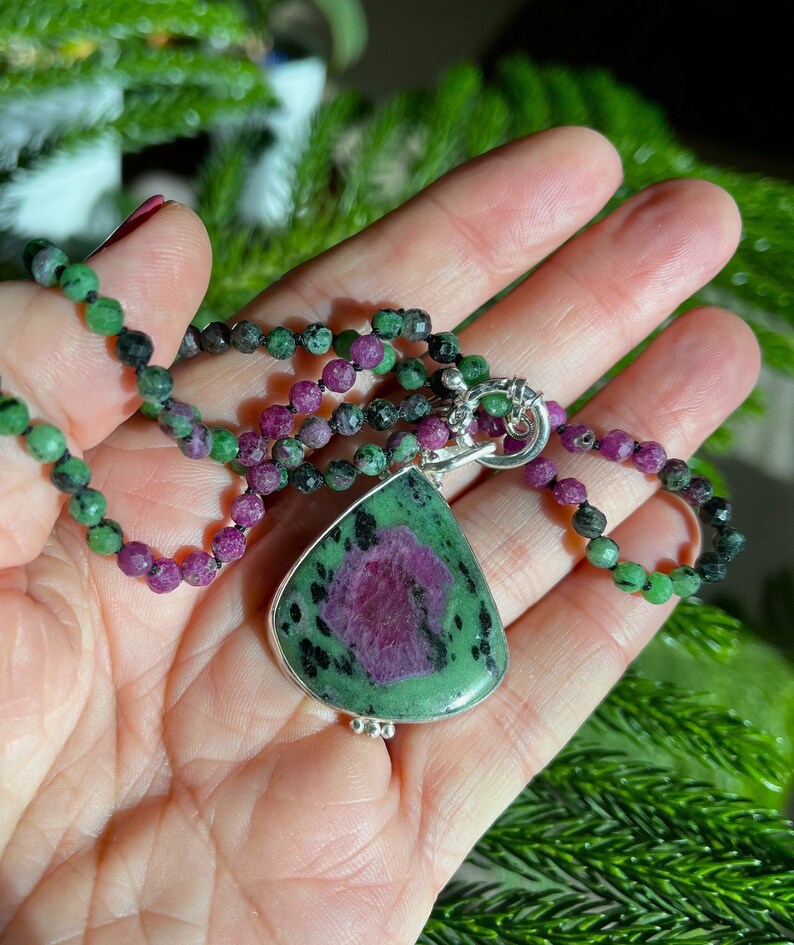 May include: A green and pink gemstone pendant necklace with a silver chain and clasp. The pendant is a teardrop shape and has a green and pink color pattern. The necklace is made of small green and pink beads.
