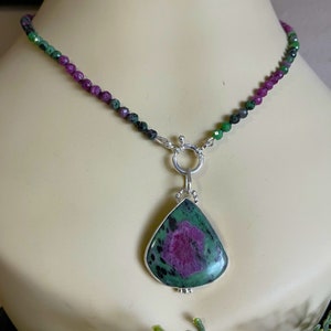 May include: A green and pink gemstone pendant necklace with a silver clasp. The pendant is a teardrop shape and is set in silver. The necklace is made of small green and pink beads.