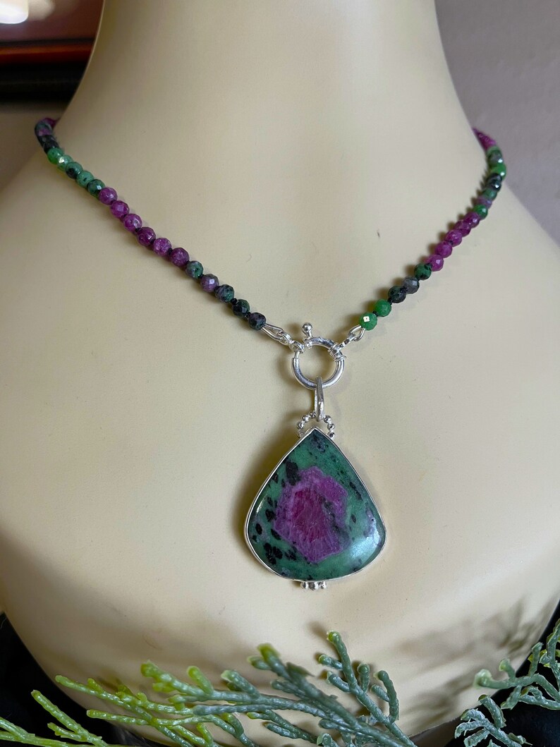 May include: A green and pink gemstone pendant necklace with a silver clasp. The pendant is a teardrop shape and is set in silver. The necklace is made of small green and pink beads.