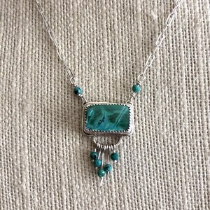 May include: A silver necklace featuring a rectangular pendant with a textured green and blue stone. The pendant is accented with small dangling turquoise-colored beads and is suspended from a silver chain with elongated oval links.