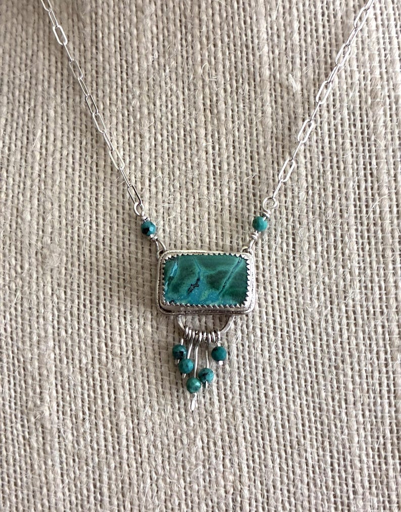May include: A silver necklace featuring a rectangular pendant with a textured green and blue stone. The pendant is accented with small dangling turquoise-colored beads and is suspended from a silver chain with elongated oval links.