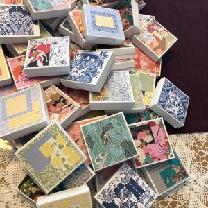 May include: A large collection of square white gift boxes, each featuring a unique decorative paper lid with various patterns including floral, geometric, and wave designs in colors such as blue, yellow, pink, green, and black.