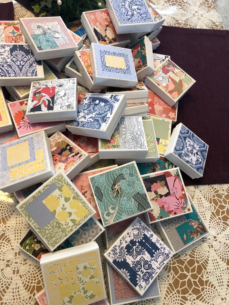 May include: A large collection of square white gift boxes, each featuring a unique decorative paper lid with various patterns including floral, geometric, and wave designs in colors such as blue, yellow, pink, green, and black.