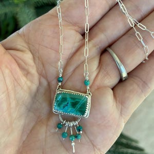 May include: A silver necklace featuring a rectangular green malachite stone set in a serrated bezel. The pendant includes a silver arch with five dangling turquoise beads and is attached to a silver paperclip chain with two additional turquoise beads.