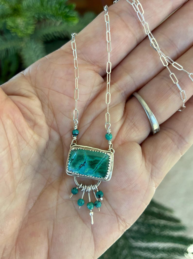 May include: A silver necklace featuring a rectangular green malachite stone set in a serrated bezel. The pendant includes a silver arch with five dangling turquoise beads and is attached to a silver paperclip chain with two additional turquoise beads.
