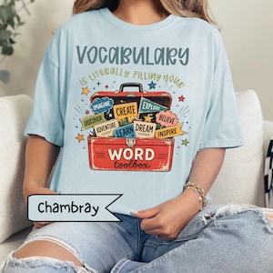 May include: A light blue t-shirt featuring a graphic of a red toolbox filled with colorful speech bubbles and banners. The text reads VOCABULARY IS LITERALLY FILLING YOUR WORD toolbox, with words like IMAGINE, CREATE, EXPLORE, DISCOVER, ADVENTURE, LEARN, DREAM, BELIEVE, and INSPIRE.