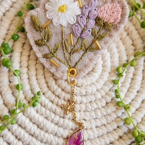 Peut inclure: Broche brodée en forme de cœur avec une marguerite blanche, de la lavande et une fleur rose. La broche a des perles dorées et un charm en forme d'abeille dorée avec un pendentif en forme de goutte rempli de fleurs roses. Sur fond beige.