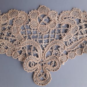May include: Close-up of a beige lace trim with an intricate floral and geometric design. The lace features a delicate, openwork pattern with scalloped edges and a symmetrical arrangement of floral motifs. The trim is likely used for decorative purposes.
