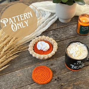 May include: A crochet pumpkin pie coaster with a white crocheted filling. The coaster is on a wooden table with a mug, a candle, and a bunch of wheat.