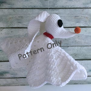 Pode incluir: Brinquedo amigurumi de crochê branco do Zero, o cão fantasma do O Estranho Mundo de Jack, com uma coleira vermelha e um nariz laranja. O brinquedo está preso a um cobertor de crochê branco com um padrão de renda. O texto "Pattern Only" é visível no cobertor.