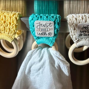 Puede incluir: Tres porta-toallas de cocina de crochet con anillos de madera. El porta-toallas del centro es turquesa con un parche cuadrado blanco que dice "Just Roll With It". El porta-toallas de la izquierda es amarillo y el porta-toallas de la derecha es blanco.