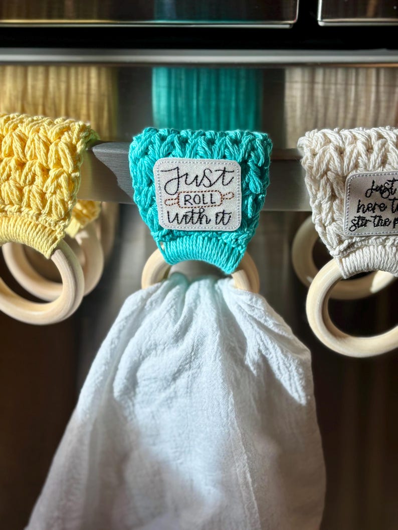 Puede incluir: Tres porta-toallas de cocina de crochet con anillos de madera. El porta-toallas del centro es turquesa con un parche cuadrado blanco que dice "Just Roll With It". El porta-toallas de la izquierda es amarillo y el porta-toallas de la derecha es blanco.