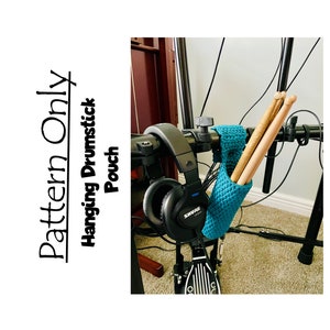 May include: A teal crochet hanging pouch for drumsticks, attached to a black metal stand. The pouch is holding three wooden drumsticks. The text "Pattern Only" is written vertically on the left side of the image. The text "Hanging Drumstick Pouch" is written vertically on the left side of the image.