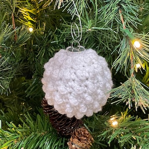 May include: White crocheted Christmas ornament hanging on a green Christmas tree. The ornament is round and has a textured surface.