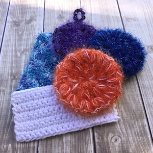 以下が含まれることがあります： 白の長方形のウォッシュクロスと、青、紫、オレンジの色合いの丸いスクラビーなど、手作りのかぎ針編みアイテムのコレクション。 スクラビーは質感のあるふわふわした外観で、木製の表面に配置されています。