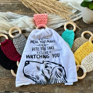 Puede incluir: Una toalla de cocina blanca con una ilustraci&oacute;n en blanco y negro de la cabeza de un perro y el texto "EVERY... MEAL YOU MAKE...EVERY... BITE YOU TAKE I'LL BE WATCHING YOU". La toalla est&aacute; colgada de un anillo de madera con un lazo de crochet rosa. Hay varios otros anillos de madera con lazos de crochet en varios colores alrededor de la toalla.