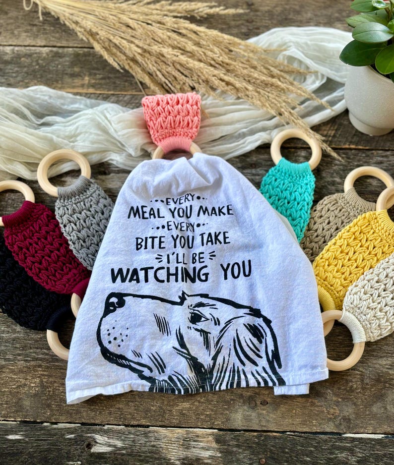 Puede incluir: Una toalla de cocina blanca con una ilustraci&oacute;n en blanco y negro de la cabeza de un perro y el texto "EVERY... MEAL YOU MAKE...EVERY... BITE YOU TAKE I'LL BE WATCHING YOU". La toalla est&aacute; colgada de un anillo de madera con un lazo de crochet rosa. Hay varios otros anillos de madera con lazos de crochet en varios colores alrededor de la toalla.