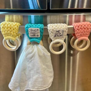 Puede incluir: Cuatro porta toallas de ganchillo con anillos de madera, colgados en un refrigerador de acero inoxidable. Los porta toallas tienen diferentes colores y lemas: "Just Roll With It", "Just Here To Stir The Pot", y dos m&aacute;s.