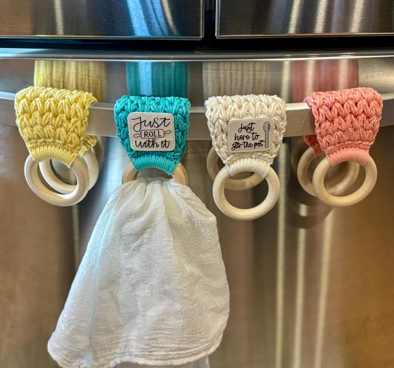 Puede incluir: Cuatro porta toallas de ganchillo con anillos de madera, colgados en un refrigerador de acero inoxidable. Los porta toallas tienen diferentes colores y lemas: "Just Roll With It", "Just Here To Stir The Pot", y dos m&aacute;s.