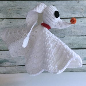 May include: White crocheted blanket with a plush, white crocheted dog head attached. The dog has black eyes, a red collar, and an orange nose.