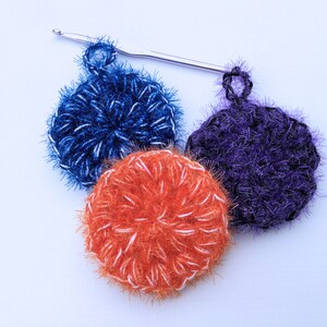May include: Three crocheted dishcloths in blue, orange, and purple. The dishcloths are made with a fluffy yarn and have a white accent color. A crochet hook is in the background.