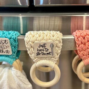 Puede incluir: Tres porta-toallas de cocina de crochet con anillos de madera. El porta-toallas de la izquierda es turquesa y dice "Just roll with it". El porta-toallas del medio es crema y dice "Just here to stir the pot". El porta-toallas de la derecha es coral.