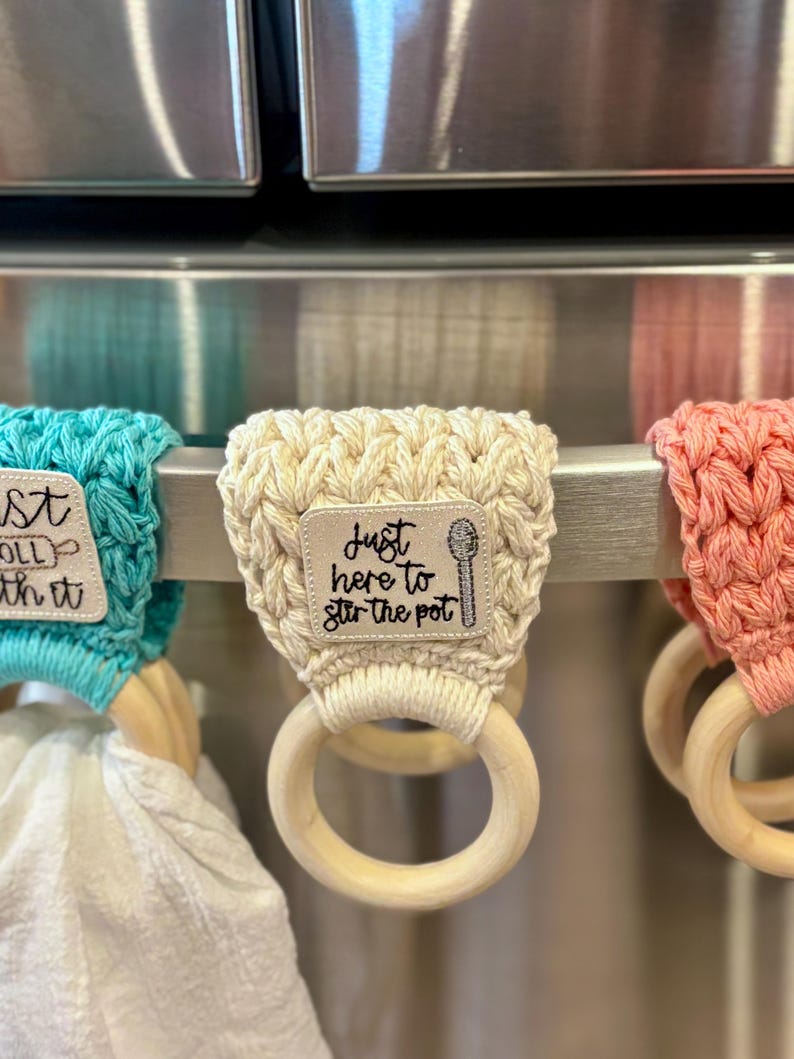 Puede incluir: Tres porta-toallas de cocina de crochet con anillos de madera. El porta-toallas de la izquierda es turquesa y dice "Just roll with it". El porta-toallas del medio es crema y dice "Just here to stir the pot". El porta-toallas de la derecha es coral.