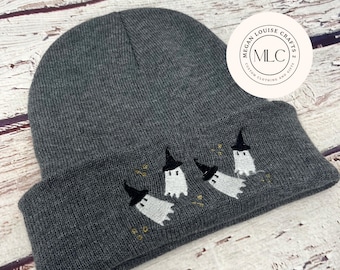 Puede incluir: Un gorro gris oscuro con un diseño bordado de tres fantasmas blancos con sombreros de bruja negros. El gorro tiene el texto "Megan Louise Crafts 2 Custom Clothing and Gifts" en un círculo blanco.