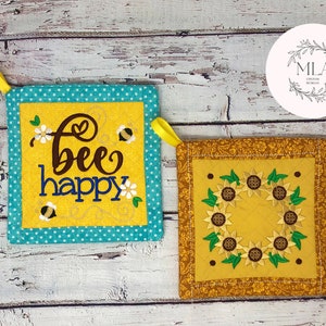 Puede incluir: Dos agarraderas de olla amarillas y marrones con diseños bordados. Una agarradera de olla presenta una abeja y el texto "bee happy". La otra agarradera de olla presenta una corona de girasoles.