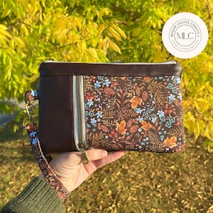 May include: A brown clutch with a floral pattern featuring blue, orange, and brown leaves and flowers. The clutch has a brown leather-like exterior, a zipper, and a wrist strap with a matching floral design. The text "Megan Louise Crafts" is on the upper right.