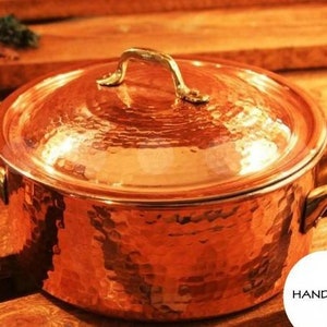 Puede incluir: Una olla de cobre martillada con tapa y asas de latón. La olla está sobre una superficie de madera. La palabra "HANDMADE" es visible en la esquina inferior derecha de la imagen.