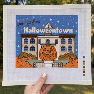 Könnte beinhalten: Eine Stickvorlage mit einem Gebäude und dem Text greetings from Halloweentown. Das Design zeigt einen großen Kürbiskopf im Vordergrund, kleinere Kürbisse, einen blauen Sternenhimmel und eine Farbpalette am rechten Rand der Leinwand.