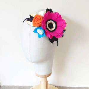 May include: A headband with a pink, orange, white, and blue felt flower arrangement. The headband is black with a gold stripe.