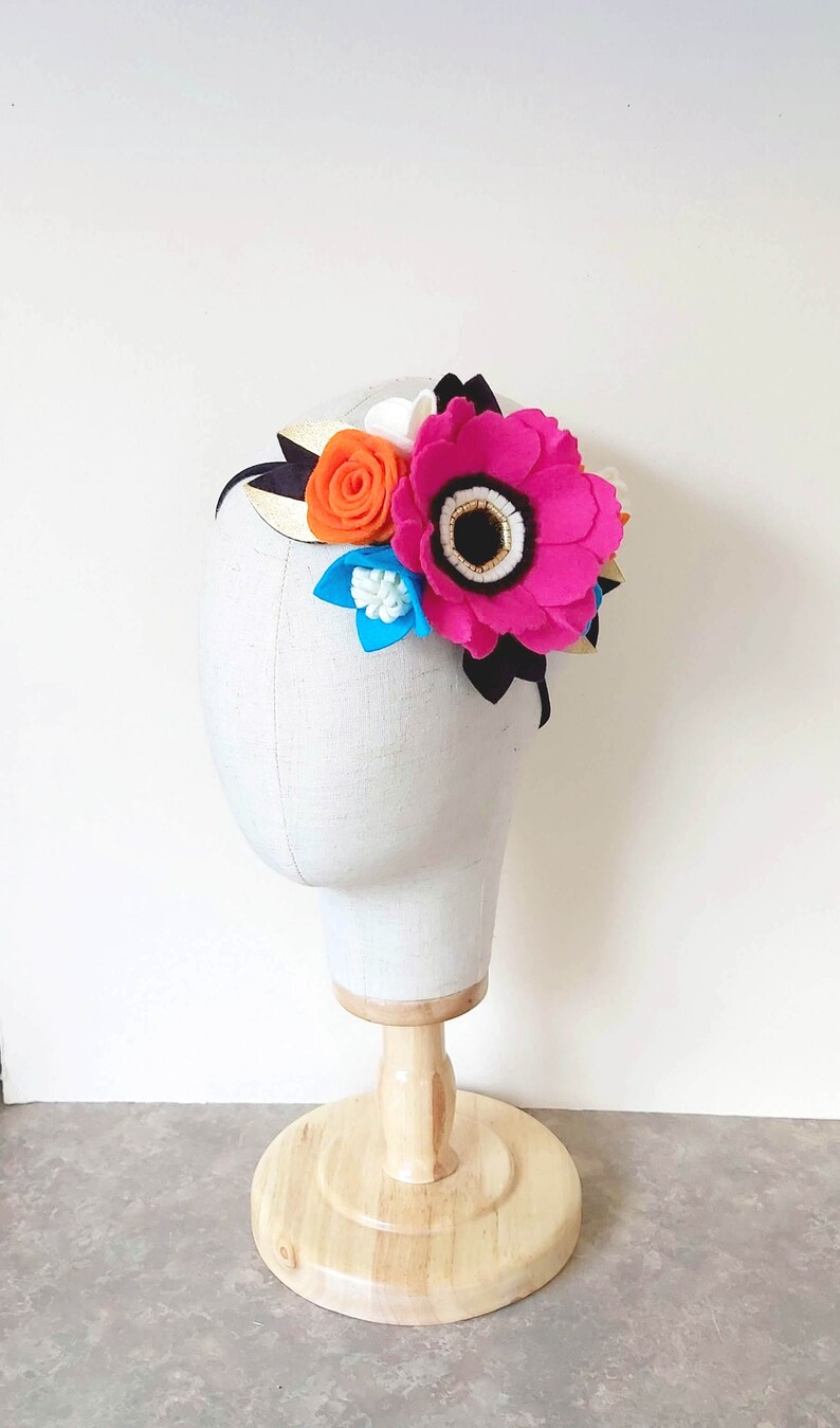 May include: A headband with a pink, orange, white, and blue felt flower arrangement. The headband is black with a gold stripe.