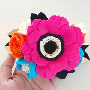 May include: A felt flower hair clip with a large pink flower, a smaller orange flower, and a blue flower. The flowers have white centers and are accented with gold beads.