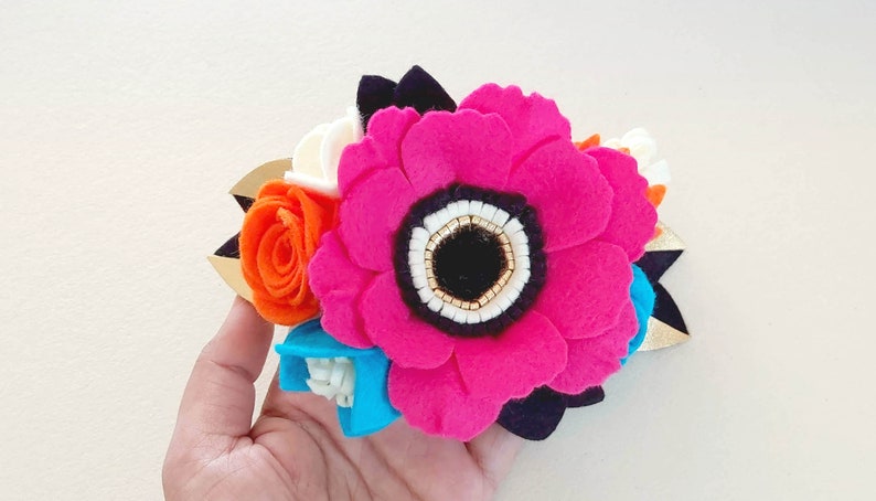 May include: A felt flower hair clip with a large pink flower, a smaller orange flower, and a blue flower. The flowers have white centers and are accented with gold beads.