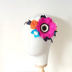 May include: A pink felt flower headband with a black felt center and white felt accents. The headband also features a smaller orange felt flower and a blue felt flower.