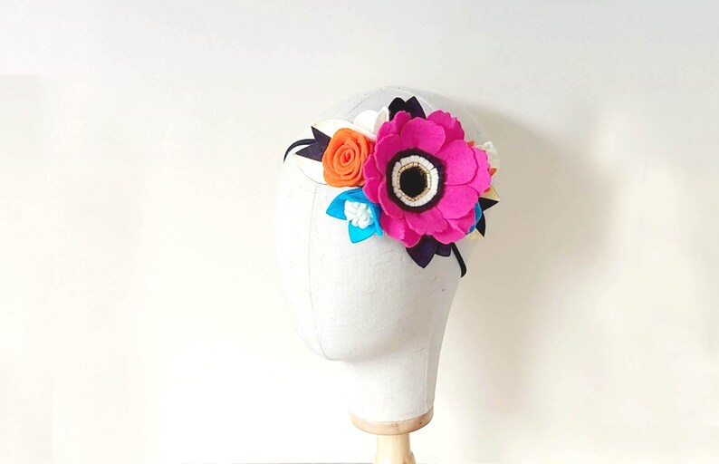 May include: A pink felt flower headband with a black felt center and white felt accents. The headband also features a smaller orange felt flower and a blue felt flower.