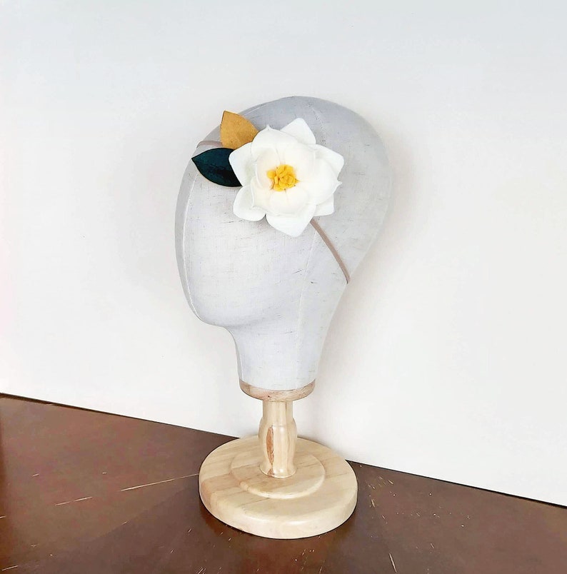 May include: A white felt flower headband with a yellow center and green leaves. The headband is on a white mannequin head.