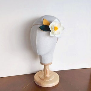 May include: A white felt flower headband with yellow and green felt leaves. The headband is on a white mannequin head.