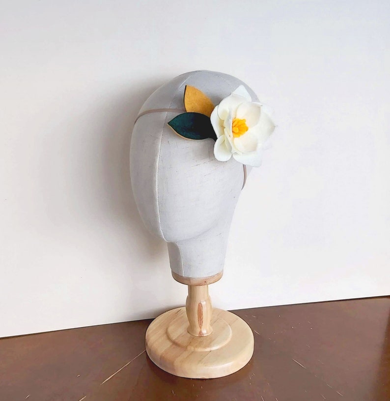 May include: A white felt flower headband with yellow and green felt leaves. The headband is on a white mannequin head.