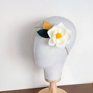 May include: A white felt flower headband with yellow and green felt leaves. The headband is on a white mannequin head.