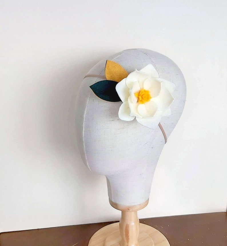 May include: A white felt flower headband with yellow and green felt leaves. The headband is on a white mannequin head.