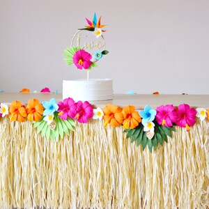 May include: A party table decorated with a grass skirt and a colourful floral garland. A white cake is topped with a gold "two" cake topper and tropical flowers. The table is set for a luau-themed birthday celebration.