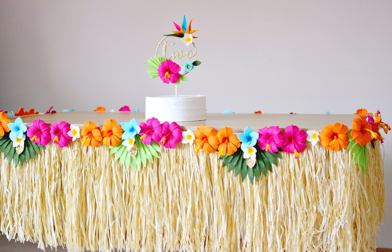 May include: A party table decorated with a grass skirt and a colourful floral garland. A white cake is topped with a gold "two" cake topper and tropical flowers. The table is set for a luau-themed birthday celebration.