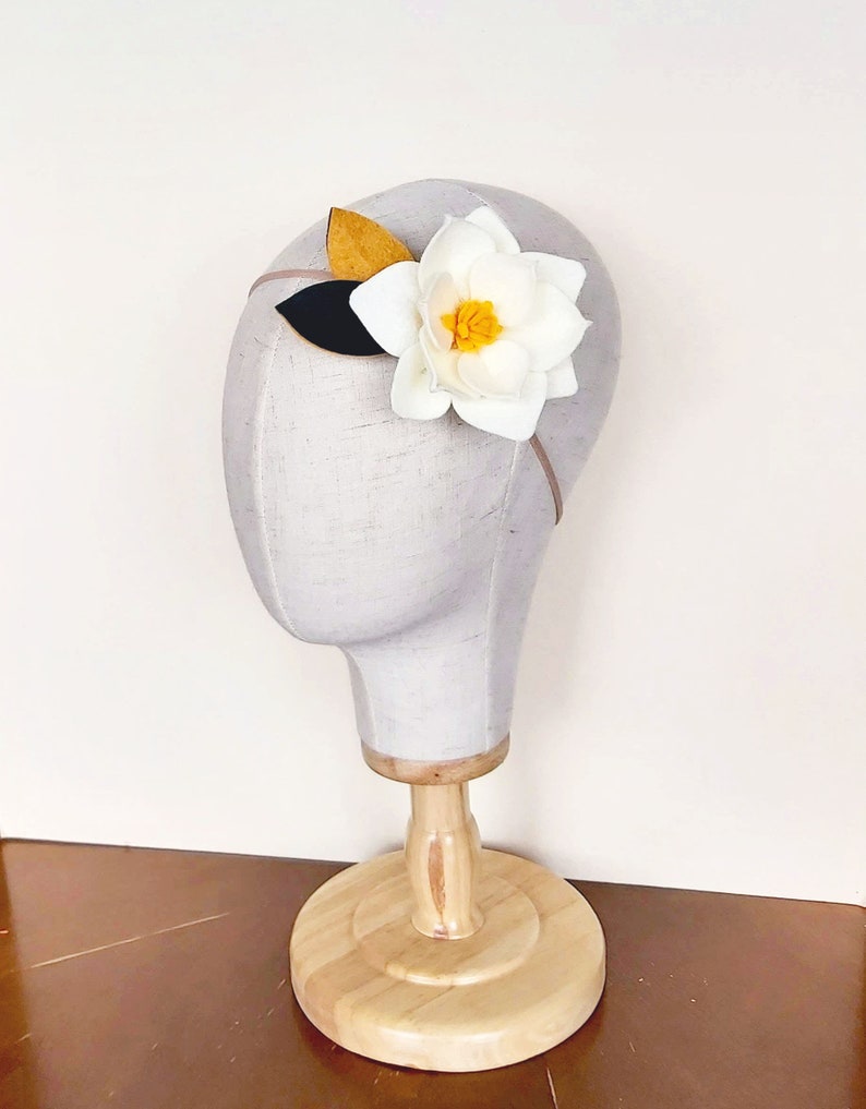 May include: A white felt flower with a yellow center and a brown felt leaf on a headband. The headband is on a white mannequin head.